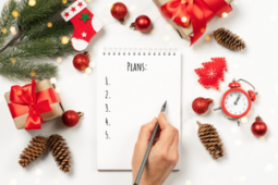 A person— surrounded by Christmas ornaments— writing their holiday plans on a flip notebook (Success Through Failure episode 326: How To Stay On Track And In Balance During The Holidays: The Ultimate Survival Guide to This Holiday Season)